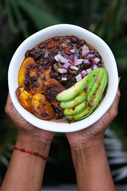 Abowl of Ghanaian beans stew with fried yellow plantains, slices of green avocado and  diced pupple onions.